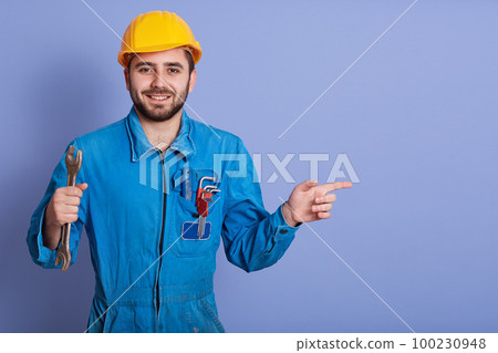 Happy and smiling bearded engineer with wrench tool in hand looking at camera and pointing aside with index finger while stands against blue studio background, dresses blueuniform and yellow helmet. Happy and smiling bearded engineer with wrench tool in hand looking at camera and pointing aside with index finger while stands against blue studio background, dresses blueuniform and yellow helmet. 100230948
