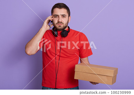 Close up portrait of serious bearded delivery man wearing red casual t shirt, talking via mobile phone with client, holding carton box in hands isolated over lilac background. Delivery concept. Close up portrait of serious bearded delivery man wearing red casual t shirt, talking via mobile phone with client, holding carton box in hands isolated over lilac background. Delivery concept. 100230999