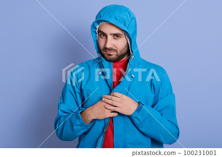 Studio shot of young attractive bearded guy wearing red shirt and blue waterproof raincoat, looking directly ar camera, posing in hood, keeping hands on zipper, male looks serious and confident. 100231001