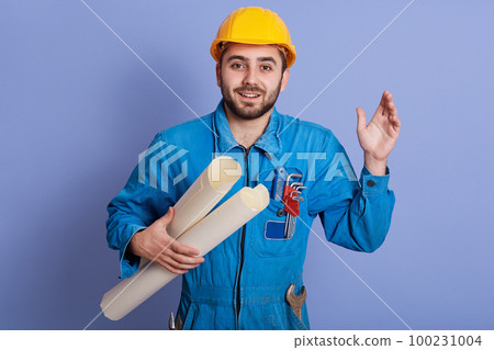 Portrait of young modern architect carrying drawings isolated over blue background, looks astonished, keeps one hand raised, posing with little bit opened mouth, dresses uniform and hard hat. 100231004