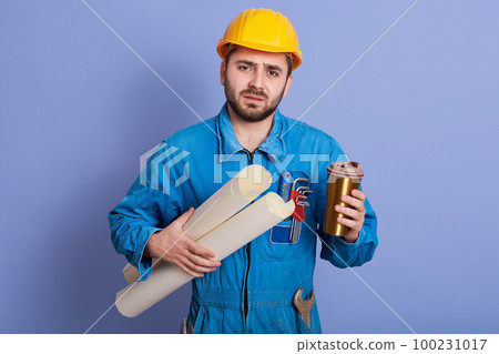 Tired engineer or foreman in protective yellow helmet and blue workwear holding blueprint in hands and enjoying hot baverage from thermo mug, looks at camera, posing isolated over studio background. 100231017