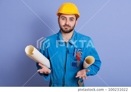 Engineer men with beard, wearing yellow safety helmet and blue uniform, handsome architect holding blueprint in hands, stands and makes helpless gesture with his palms isolated over studio background. Engineer men with beard, wearing yellow safety helmet and blue uniform, handsome architect holding blueprint in hands, stands and makes helpless gesture with his palms isolated over studio background. 100231022