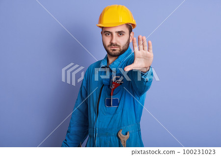 Young bearded handsome workman with yellow helmet and uniform making stop gesture with his hand denying situation that thinks wrong, posing with serious expression isolated over blue background. Young bearded handsome workman with yellow helmet and uniform making stop gesture with his hand denying situation that thinks wrong, posing with serious expression isolated over blue background. 100231023