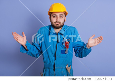 Close up portrait surprised bearded builder dresses helmet and uniform, spreading his hands to sides, squeezing his shoulders, posing isolated over blue background, guy with wrench tools working. 100231024