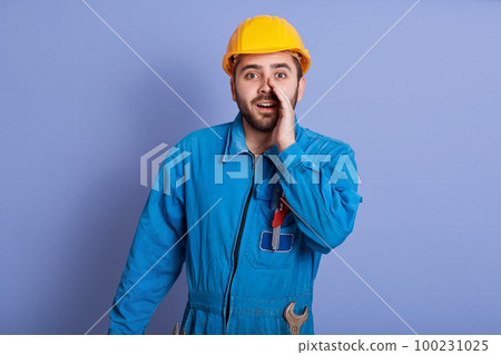 Close up portrait of bearded handsome male constructor yelling something and keeping hands near his opened mouth, foreman wearing uniform and yellow hard hat, posing isolated over blue background. 100231025