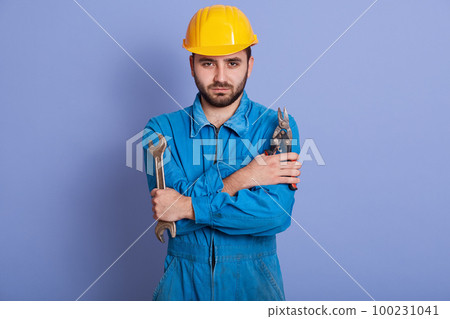 Indoor portrait of confident handsome bearded worker standing isolated over blue background in studio, holding pliers and wrench in both hands, being ready to work. People and engineering concept. 100231041