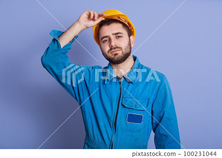 Studio shot of man wearing blue uniform and yellow building helmet looking at camera, keeping hand on his hard hat, handsome guy with dark beard, looks tired after hard work. Building concept. 100231044