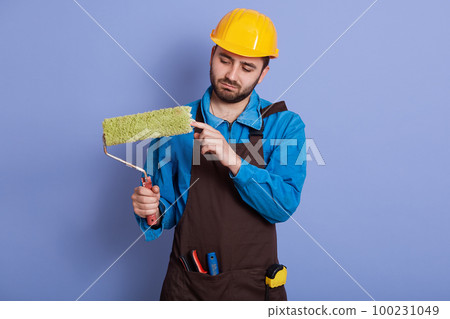 Horizontal picture of attentive hard working young repairman holding roller in one hand, wearing blue overall, brown apron and yellow helmet, having equipment. People and working process concept. Horizontal picture of attentive hard working young repairman holding roller in one hand, wearing blue overall, brown apron and yellow helmet, having equipment. People and working process concept. 100231049