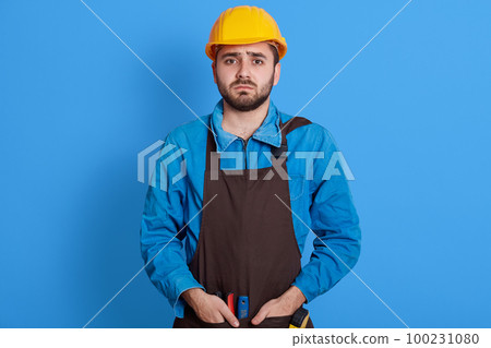 Helpless bearded engineer wearing yellow protective helmet keeping hands in pockets of apron, looks sad and upset, does not know what to do, posing isolated over blue background. Helpless bearded engineer wearing yellow protective helmet keeping hands in pockets of apron, looks sad and upset, does not know what to do, posing isolated over blue background. 100231080
