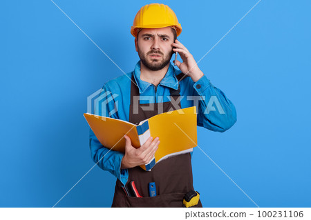 Young European bearded man working as architect or foreman, solving problems on building, holds paper folder, talking phone, wearing apron, helmet, and blue uniform. Young European bearded man working as architect or foreman, solving problems on building, holds paper folder, talking phone, wearing apron, helmet, and blue uniform. 100231106