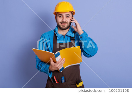 Close up portrait of young attractive bearded builder having conversation via phone and holding paper folder in hands, dresses blue shirt, brown apron, yellow helmet. Buildingand engineering concept. 100231108