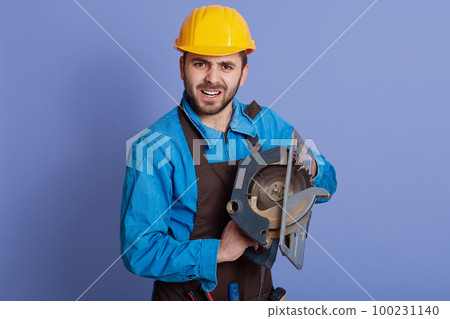 Man holding circular in hands, has angry facial expression, bearded working man wearing apron and yellow protective helmet, posing isolated over blue background. 100231140