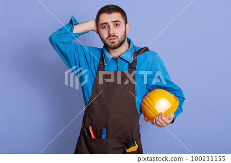 Horizontal indoor picture of confused disappointed builder having no idea what to do, losing job, having no projects because of coronavirus, looking directly at camera, holding helmet in one hand. 100231155