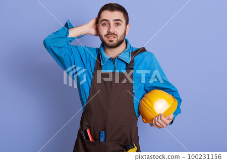 Picture of surprised emotional builder putting hand on head, holding yellow helmet, wearing overall and brown apron, looking directly at camera, having tools, being involved in work. Work concept. 100231156