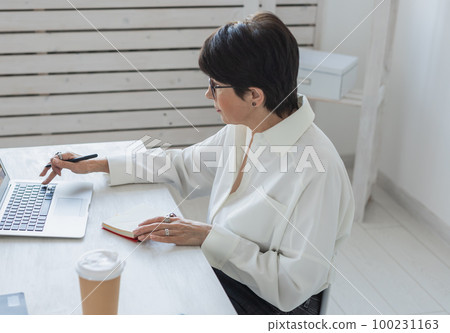 Middle-aged beautiful woman smiling while working with laptop in office - business and manager works concept 100231163