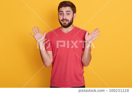 Close up portrait of young bearded man dresses casual red shirt posing isolated over yellow wall background. People emotions, lifestyle concept. Guy showing palms, rising hands up, looks astonished. 100231228