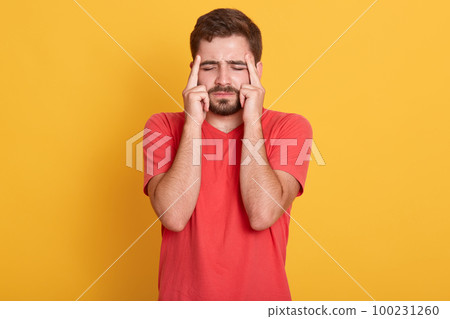 Portrait of young bearded man isolated over yellow background, suffering from severe headache, pressing fingers to temples, closing eyes to relieve pain, guy wears casual clothing. Health care concept Portrait of young bearded man isolated over yellow background, suffering from severe headache, pressing fingers to temples, closing eyes to relieve pain, guy wears casual clothing. Health care concept 100231260