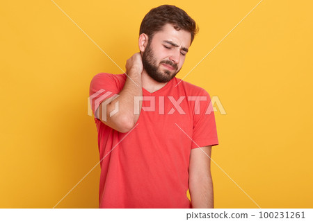 Image of attractive bearded male feeling pain in neck, frustrated young man wearing red casual t shirt, touching his neck and expressing negativity while standing against yellow studio background. 100231261