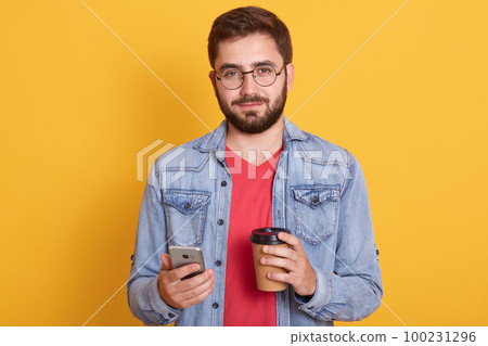 Photo of confident good looking young man holding paper cup with coffee and smartphone, wearing denim jacket, eyewear and t shirt, having lunch, chatting online. People and technology concept. Photo of confident good looking young man holding paper cup with coffee and smartphone, wearing denim jacket, eyewear and t shirt, having lunch, chatting online. People and technology concept. 100231296