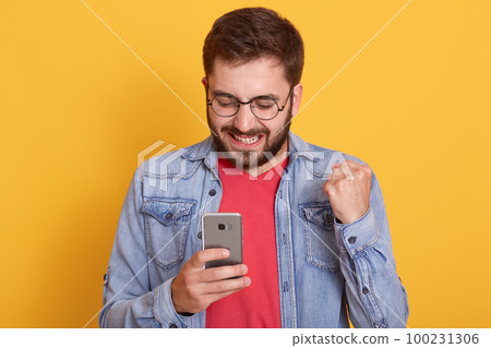 Portrait of smiling happy man wearing denim jacket and red shirt, clenching fist and holding smart phone in hands, male sees something excited on his device's screen. Portrait of smiling happy man wearing denim jacket and red shirt, clenching fist and holding smart phone in hands, male sees something excited on his device's screen. 100231306