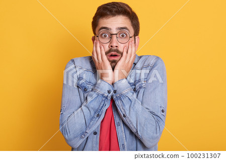 Portrait of emotional shocked scared guy opening eyes and mouth widely, putting hands to face, having impressed facial expression, wearing denim jacket and red t shirt. People and emotions concept. Portrait of emotional shocked scared guy opening eyes and mouth widely, putting hands to face, having impressed facial expression, wearing denim jacket and red t shirt. People and emotions concept. 100231307