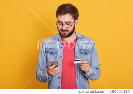 Studio shot of young european bearded man holding credit card and smartphone, standing with calm facial expression over yellow background, spending money from his account. Online shopping concept. 100231315