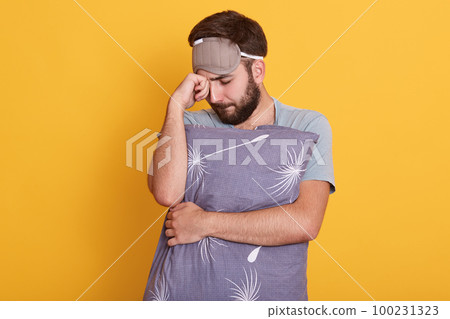 Studio shot of youngbearded man with dark hair, keeping his fist near forehead, keeps eyes closed, thinks about something important or having problems, male embracing grey pillow against yellow wall. 100231323
