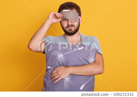 Portrait of bearded man in dressing gown and mask to sleep on his head, wants to continue sleeping, do not wake up in morning, isolated over yellow background, unshaven sleepy guy hugging pillow, 100231332