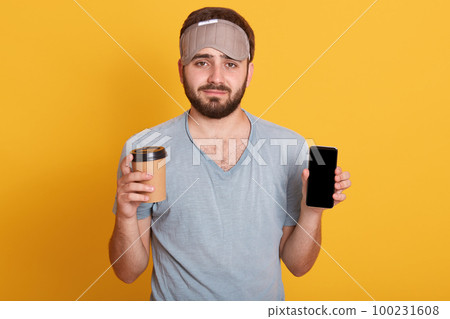 Portrait of upset handsome bearded young hipster man in gray t shirt and blindfold standing with coffee in one hand and phone with blank screen in another, isolated over pink studio background. 100231608