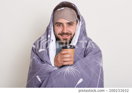 Indoor shot of positive unshaven cheerful young European man posing isoated over white studio wall with plaid on his shoulders and blindfold on his forehead, enjoying coffee or tea. Morning concept. Indoor shot of positive unshaven cheerful young European man posing isoated over white studio wall with plaid on his shoulders and blindfold on his forehead, enjoying coffee or tea. Morning concept. 100231632