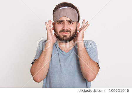 Portrait of irritated angry young man having beard, looking directly at camera, covering his ears with fingers, wearing t shirt and sleeping mask, posing isolated over white background in studio. Portrait of irritated angry young man having beard, looking directly at camera, covering his ears with fingers, wearing t shirt and sleeping mask, posing isolated over white background in studio. 100231646