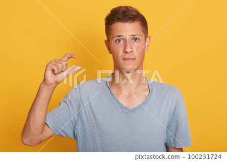 Indoor studio shot of disappointed cute young man looking directly at camera, posing in casual blue t shirt, making gesture, showing measure with two fingers, feeling upset. Emotions concept. 100231724