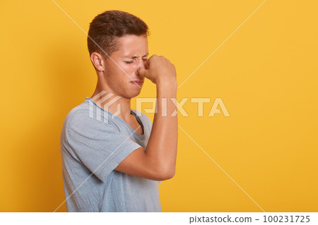 Indoor image of upset miserable young man standing in profile, closing his eyes, blocked his nose with hand, feeling unpleasant smell, wearing casual blue t shirt. Copyspace for advertisement. 100231725