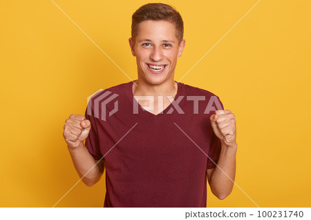 Happy positive excited young man clenching fists and screaming, attractive guy wearing maroon casual t shirt glad to hear good news, celebrating his victory or success, isolated over yellow background Happy positive excited young man clenching fists and screaming, attractive guy wearing maroon casual t shirt glad to hear good news, celebrating his victory or success, isolated over yellow background 100231740