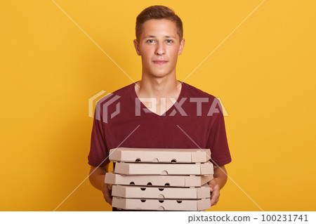 Boy wearing maroon casual t shirt delivering pizza boxes, posing isolated over yellow background, looking at camera, looks serious, young female working as delivery man, doing his work. People concept 100231741