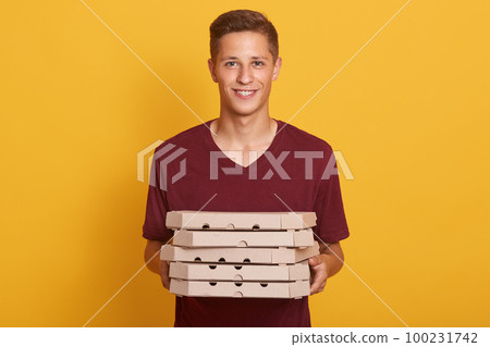Image of joyful handsome delivery man wearing casual burgundy t shirt, holding stack of pizza boxes in hands and looking directly at camera isolated over yellow studio background. Junk food concept. Image of joyful handsome delivery man wearing casual burgundy t shirt, holding stack of pizza boxes in hands and looking directly at camera isolated over yellow studio background. Junk food concept. 100231742