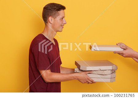 Profile of cheerful pleasant young boy holding cardboard boxes with pizza, unknown hands putting one more box, smiling sincere, standing isolated over yellow background in studio. Delivery concept Profile of cheerful pleasant young boy holding cardboard boxes with pizza, unknown hands putting one more box, smiling sincere, standing isolated over yellow background in studio. Delivery concept 100231765