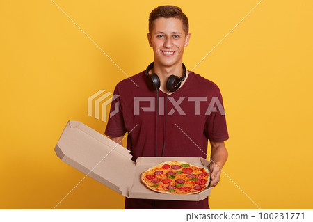 Indeoor of magnetic young boy holding opened box with pizza, having headphones around neck, wearing burgundy t shirt, male posing isolated over yellow background in studio. Delivery concept. 100231771