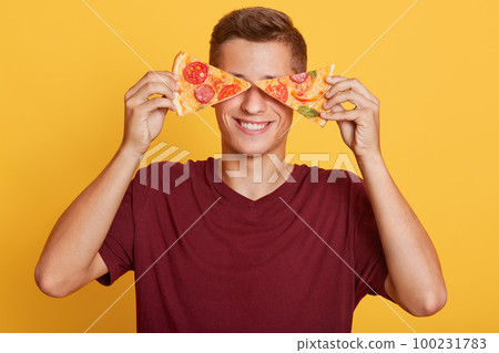 Image of cheerful handsome young boy holding two pieces of pizza in both hands, having beautiful smile, covering his eyes with pizza, being in high spirits, fastfood lover. People and food concept. Image of cheerful handsome young boy holding two pieces of pizza in both hands, having beautiful smile, covering his eyes with pizza, being in high spirits, fastfood lover. People and food concept. 100231783