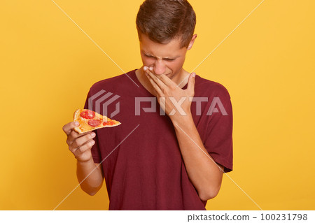 Young Caucasian student male fast food lover dresses burgundy t shirt gets his order from pizzeria, holding slice of expired pizza, feels bad smell, covers mouth with palm, stands against yellow wall. Young Caucasian student male fast food lover dresses burgundy t shirt gets his order from pizzeria, holding slice of expired pizza, feels bad smell, covers mouth with palm, stands against yellow wall. 100231798