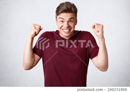 Happy joyful strong male clenches fists and teeth, dressed in casual t shirt, celebrates his victory and success, isolated over white background. Glad cheerful man youngster gestures indoor. Happy joyful strong male clenches fists and teeth, dressed in casual t shirt, celebrates his victory and success, isolated over white background. Glad cheerful man youngster gestures indoor. 100231908