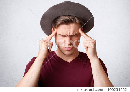 Studio shot of sullen Caucasian male frowns face, keeps fore fingers on temples, has discontent expression, poses against white background. People, facial expressions and negativity concept. Studio shot of sullen Caucasian male frowns face, keeps fore fingers on temples, has discontent expression, poses against white background. People, facial expressions and negativity concept. 100231932