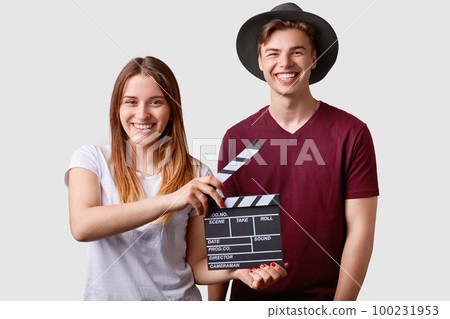 Two successful young female and male famous producers or directors hold film clapper, participate in shooting film, have joyful expressions, pose against white background. Film making concept. Two successful young female and male famous producers or directors hold film clapper, participate in shooting film, have joyful expressions, pose against white background. Film making concept. 100231953