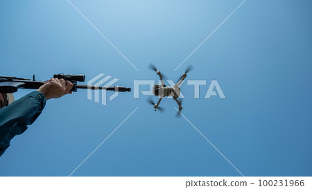 A man aims to shoot a rifle at a flying drone against a blue sky.  100231966