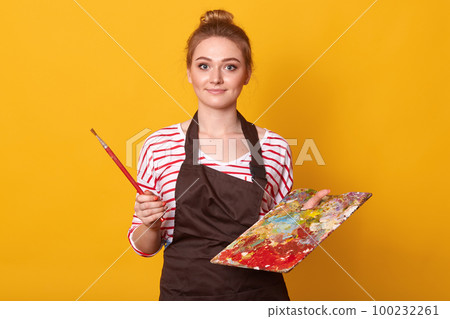Surprised emotional blond haired female posing with bunch on her head, having impressed facial expression, holding paint equipment for her hobby, isolated over bright yellow background in studio. 100232261