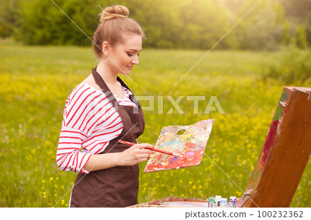 Picture of pleseant looking concentrated young Caucasian female with fair hair, artist standing in meadow with painting accessories, holding palette of colours, painting beautiful landscapes on canvas 100232362