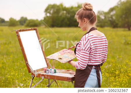 Image of female artist working with watercolor painting, posing in open air, female wearing white casual shirt with red stripes, has blonde hair, lady drawing landscapes. Creativity and art concept. Image of female artist working with watercolor painting, posing in open air, female wearing white casual shirt with red stripes, has blonde hair, lady drawing landscapes. Creativity and art concept. 100232367
