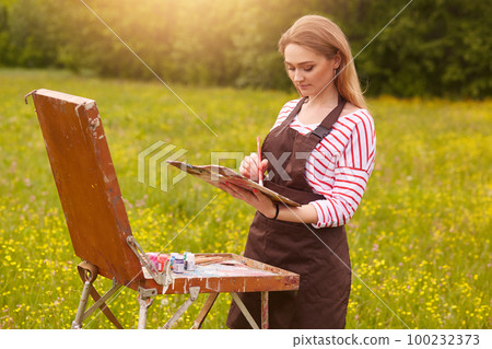 Close up portrait of beautiful girl with painting picture on easel in nature, young attractive woman involved in creativity and enjoying beautiful landscape on summer day, being happy to do she likes. 100232373