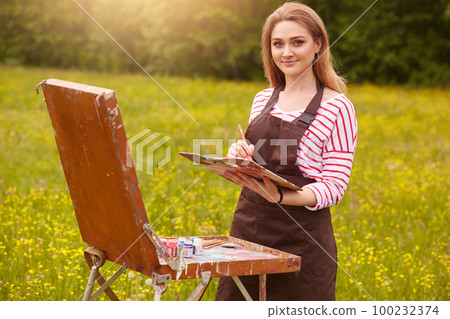 Artist paints picture in open air, holding oil paint brush in hand and palette of colours, bringing her creativity to life. Lady with charming smile looks at camera, wearing casually, draws landscape. 100232374