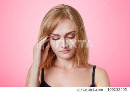 Serious pleasant looking young female model keeps hand on temples, looks pensively down, has thoughtful expression, tries to remember something, has headache after long working day, rests at home Serious pleasant looking young female model keeps hand on temples, looks pensively down, has thoughtful expression, tries to remember something, has headache after long working day, rests at home 100232405